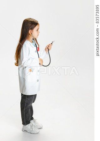 Side view. Cute beautiful little girl, child in image of doctor wearing white lab coat posing isolated on white studio background Side view. Cute beautiful little girl, child in image of doctor wearing white lab coat posing isolated on white studio background 83573895
