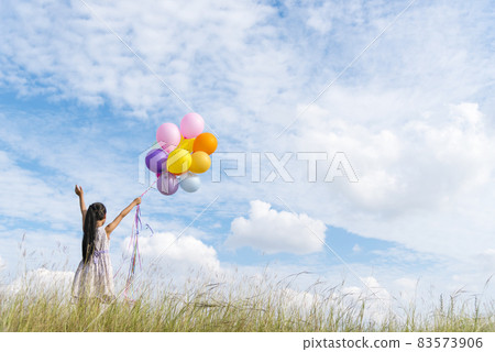 Cheerful cute girl holding balloons running on green meadow white cloud and blue sky with happiness. Hands holding vibrant air balloons play on birthday party happy times summer on sunlight outdoor 83573906