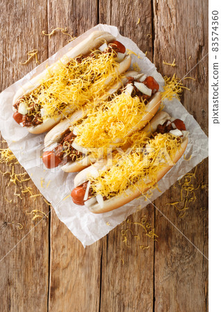 Cincinnati fast food chili hot dog with beef sausage, cheddar cheese and onions close-up. Vertical top view Cincinnati fast food chili hot dog with beef sausage, cheddar cheese and onions close-up. Vertical top view 83574360