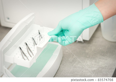 The beautician holds a tray of tweezers preparing them for disinfection with a special liquid and machine. 83574708