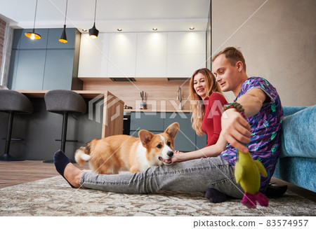 Cheeful couple sitting on carpet leaning on couch and having fun while playing with dog by soft toy. On the background kitchen furniture, loft style lamps and table with chairs. Cheeful couple sitting on carpet leaning on couch and having fun while playing with dog by soft toy. On the background kitchen furniture, loft style lamps and table with chairs. 83574957