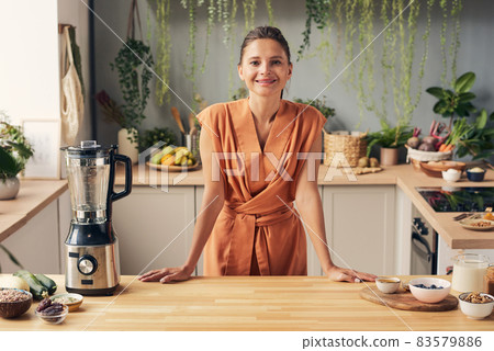 Happy young female in orange dress standing by kitchen table Happy young female in orange dress standing by kitchen table 83579886