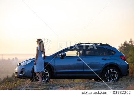 Happy young woman driver in blue dress enjoying warm summer evening standing beside her car. Travelling and vacation concept. Happy young woman driver in blue dress enjoying warm summer evening standing beside her car. Travelling and vacation concept. 83582262