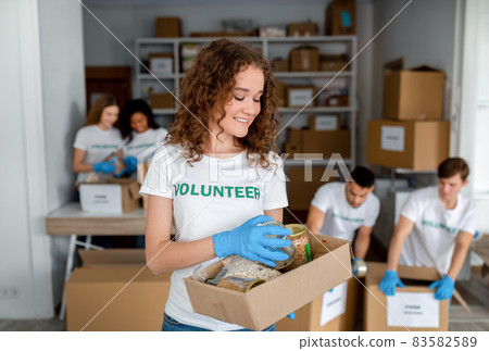 Portrait of happy female volunteer holding donations box with food and cans, diverse colleagues packing products Portrait of happy female volunteer holding donations box with food and cans, diverse colleagues packing products 83582589