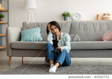Full length of unhappy Indian woman sitting on floor with joystick, losing online video game, feeling stressed at home Full length of unhappy Indian woman sitting on floor with joystick, losing online video game, feeling stressed at home 83582732