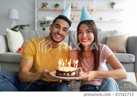 Cheery millennial multiracial couple in party hats holding birthday cake with lit candles, smiling at camera indoors 83582733