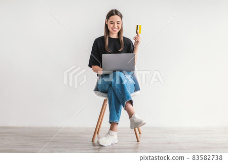 Young woman with credit card and laptop sitting on chair, buying goods on internet. Online shopping, remote banking Young woman with credit card and laptop sitting on chair, buying goods on internet. Online shopping, remote banking 83582738