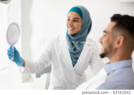 Muslim Dentist Woman Holding Mirror, Showing Teeth Treatment Result To Male Patient 83582842