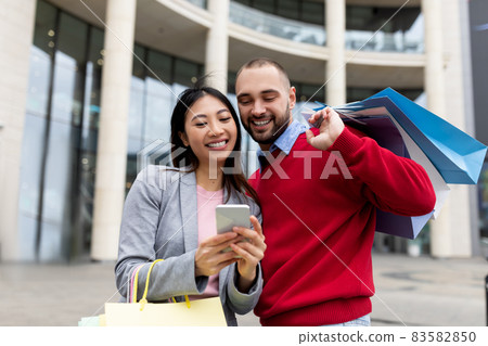 Portrait of young multiracial couple using smartphone shopping app, holding bright gift bags near city mall, outdoors 83582850