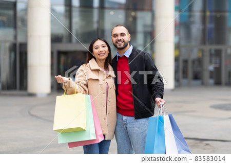 Happy international couple in casual wear posing with shopping bags, looking at camera and smiling near city mall 83583014
