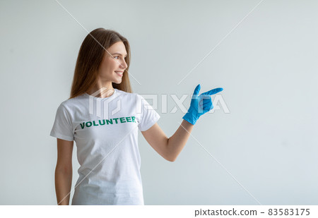Look here. Young female volunteer in gloves pointing and looking aside at free space, standing over light studio wall Look here. Young female volunteer in gloves pointing and looking aside at free space, standing over light studio wall 83583175