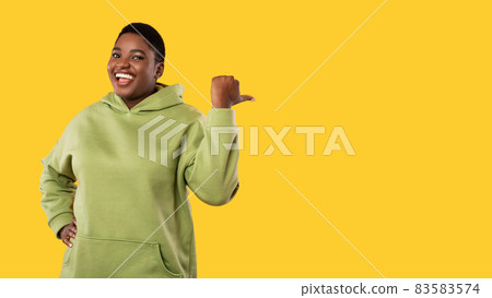 Cheerful Obese Black Woman Pointing Thumb Finger Aside, Yellow Background Cheerful Obese Black Woman Pointing Thumb Finger Aside, Yellow Background 83583574