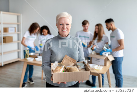 Happy senior woman holding box with donations food and smiling at camera while group of volunteers packing boxes Happy senior woman holding box with donations food and smiling at camera while group of volunteers packing boxes 83583727