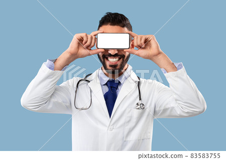 Male Doctor Holding Smartphone In Front Of Eyes, Blue Background Male Doctor Holding Smartphone In Front Of Eyes, Blue Background 83583755