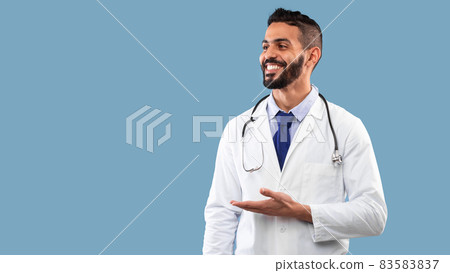 Cheerful Middle Eastern Doctor Showing Free Space Over Blue Background, Panorama Cheerful Middle Eastern Doctor Showing Free Space Over Blue Background, Panorama 83583837
