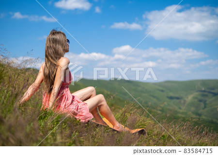 Young woman in red dress resting on green grassy field on a sunny day in summer mountains enjoying view of nature. Young woman in red dress resting on green grassy field on a sunny day in summer mountains enjoying view of nature. 83584715