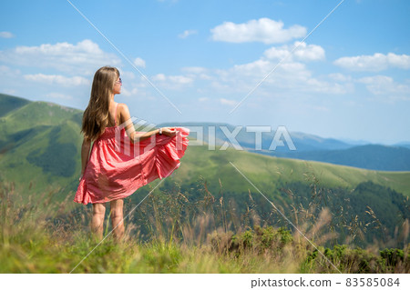 Young woman in red dress standing on grassy field on a windy day in summer mountains enjoying view of nature. 83585084