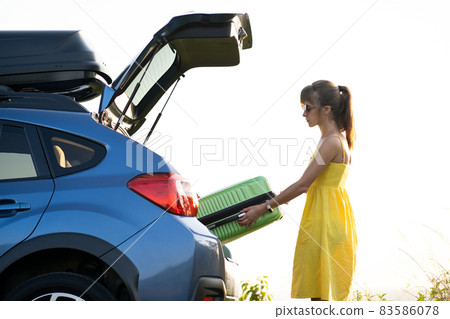 Young woman in yellow summer dress taking green suitcase from car trunk. Travel and vacations concept. 83586078