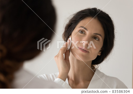 Woman looks in mirror holds cotton disc cleanse facial skin Woman looks in mirror holds cotton disc cleanse facial skin 83586304
