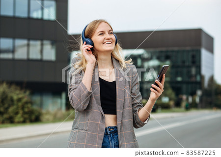Portrait of young happy blonde woman listening to music with headphones and smiling while walking on the street in the city. Music lover enjoying music. Portrait of businesswoman walking Portrait of young happy blonde woman listening to music with headphones and smiling while walking on the street in the city. Music lover enjoying music. Portrait of businesswoman walking 83587225
