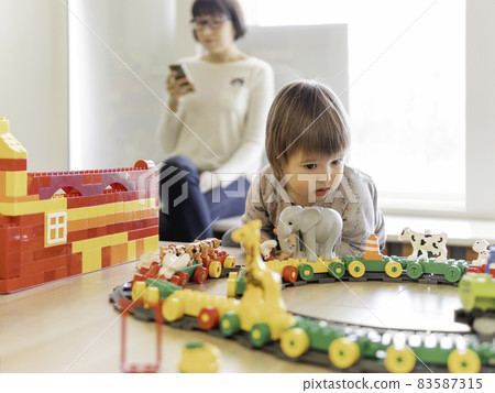 Toddler plays with colorful toy blocks while his mother or babysitter texting in smartphone. Little boy stares on toy constructor. Interior of kindergarten or nursery. 83587315