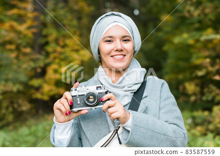 Young Arab Woman wearing hijab headscarf photographing with a smartphone in park. Modern muslim girl Young Arab Woman wearing hijab headscarf photographing with a smartphone in park. Modern muslim girl 83587559
