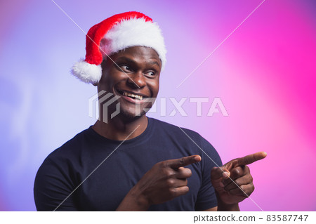 Portrait of positive african american smiling man in santa hat and casual t-shirt on colourful background, christmas time. Winter holidays and emotional people concept. Portrait of positive african american smiling man in santa hat and casual t-shirt on colourful background, christmas time. Winter holidays and emotional people concept. 83587747