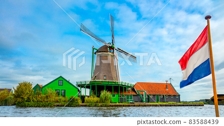 Wind mill, old house and the flag of the Netherlands are popular Dutch icons. Landscape, panorama. Amsterdam, Holland, Netherlands. 83588439