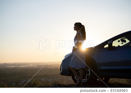 Happy young woman driver in blue dress enjoying warm summer evening standing beside her car. Travelling and vacation concept. Happy young woman driver in blue dress enjoying warm summer evening standing beside her car. Travelling and vacation concept. 83588965