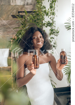 Pretty dark-skinned woman civered with towel choosing body lotion Pretty dark-skinned woman civered with towel choosing body lotion 83594012
