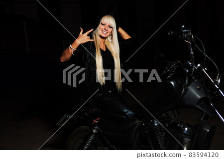Blond haired biker girl is sitting on motorbike in night anf gesturing - shows mano cornuta or devils horns sign Blond haired biker girl is sitting on motorbike in night anf gesturing - shows mano cornuta or devils horns sign 83594120
