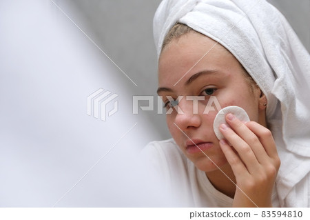 A teenage girl wiping her face with a cotton pad near the mirror. Facial skin care in adolescence. 83594810