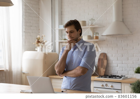 Man standing in kitchen staring at laptop thinks over task 83601156