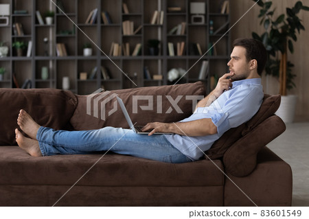 Pensive young man using computer, lying on sofa. Pensive young man using computer, lying on sofa. 83601549