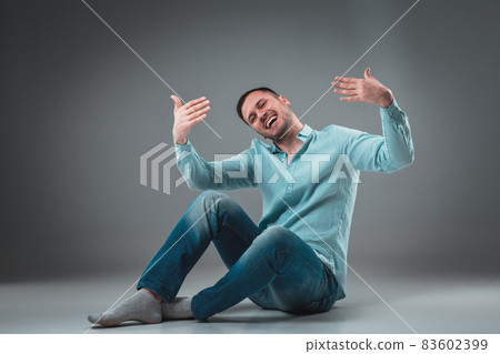 Appealing casual young man sitting on the floor, looking to the camera smiling Appealing casual young man sitting on the floor, looking to the camera smiling 83602399