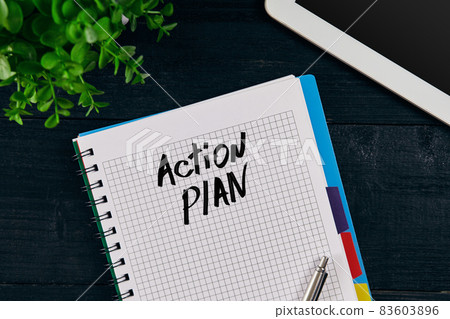 Top view of open notebook written with ACTION PLAN inscription. Green flower, tablet, pen and notebook on wooden background. 83603896