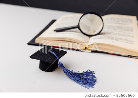 Magnifying glass on open book and graduation cap on white table. Education concept Magnifying glass on open book and graduation cap on white table. Education concept 83605666