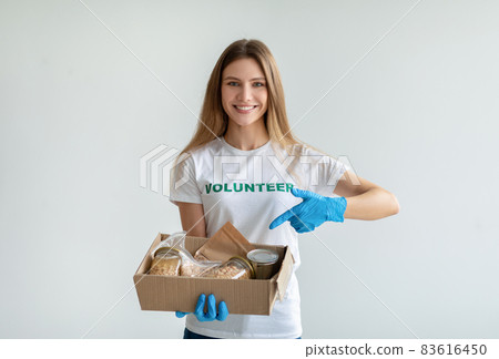 Happy female volunteer holding and pointing at box with donations, smiling at camera, standing over light background 83616450