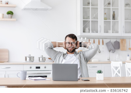 Smiling adult european businessman in glasses resting from work and looking at laptop at kitchen Smiling adult european businessman in glasses resting from work and looking at laptop at kitchen 83616742