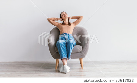 Peaceful young woman sitting in armchair, holding hands behind head, relaxing, having break against white studio wall Peaceful young woman sitting in armchair, holding hands behind head, relaxing, having break against white studio wall 83617028
