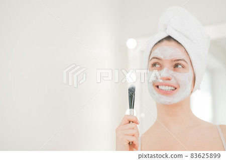 Beautiful caucasian woman with a towel on her hair applies a clay face mask. Taking care of beauty at home Beautiful caucasian woman with a towel on her hair applies a clay face mask. Taking care of beauty at home 83625899