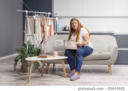 Wondering looking at the window young fat girl working on laptop sitting on the sofa at home with the kitchen on background. Working distantly from home using laptop 83633692
