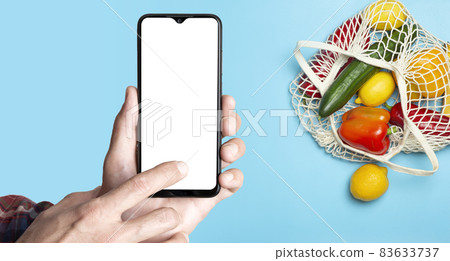 Grocery online background concept. A hand with a smartphone and eco-friendly bag with vegetables and fruit from the supermarket. Order, grocery and delivery food from eco-market. 83633737