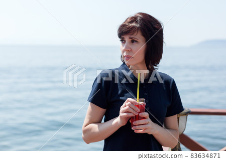 A brunette woman in a cafe on the pier is standing with a glass of juice waiting for the ship 83634871