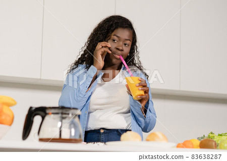 Afro Woman Talks on Mobile And Drinks an Orange Juice From a Straw in Her Kitchen. Close-up. White Background Afro Woman Talks on Mobile And Drinks an Orange Juice From a Straw in Her Kitchen. Close-up. White Background 83638287