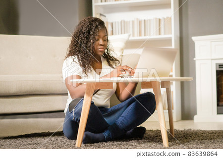 Pretty Afro Girl With Folded Legs Sitting on the Carpet and Typing on her Computer. African Girl Looks Through the List of Films on Her Laptop. Close-up. Home Background Pretty Afro Girl With Folded Legs Sitting on the Carpet and Typing on her Computer. African Girl Looks Through the List of Films on Her Laptop. Close-up. Home Background 83639864