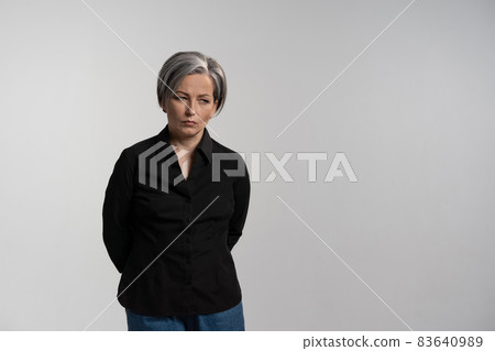 Distressed or disappointed mature grey haired woman looking down. Pretty mid aged grey haired woman in black shirt isolated on grey background. Human emotions, facial expression concept Distressed or disappointed mature grey haired woman looking down. Pretty mid aged grey haired woman in black shirt isolated on grey background. Human emotions, facial expression concept 83640989