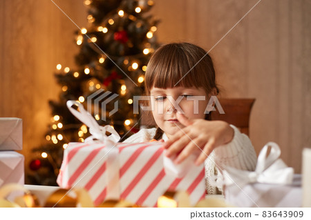 Portrait of charming adorable little girl packing present boxes for Christmas, dark haired female child with curious expression preparing for winter holidays. 83643909