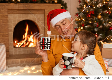 Female kid ans her caring grandfather drinking hot tea from mugs while sitting on floor in festive living room, enjoying to spend winter holidays together. Female kid ans her caring grandfather drinking hot tea from mugs while sitting on floor in festive living room, enjoying to spend winter holidays together. 83643981
