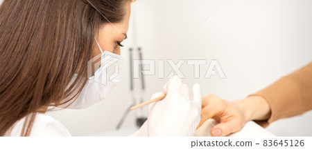 Manicure master in protective face mask and white gloves apply polish to female fingernails. Manicure master in protective face mask and white gloves apply polish to female fingernails. 83645126